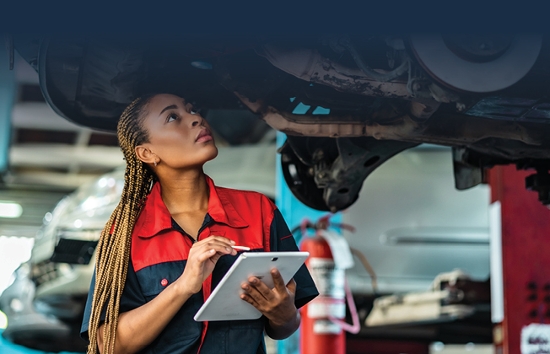 Una mujer mecánica usando un overol en su trabajo en un taller de reparación de autos. Está mirando hacia arriba hacia el chasis de un vehículo de flota elevado en una grúa, consultando su tableta en una mano y marcando la lista de verificación de las tareas de mantenimiento con el lápiz óptico en la otra mano.